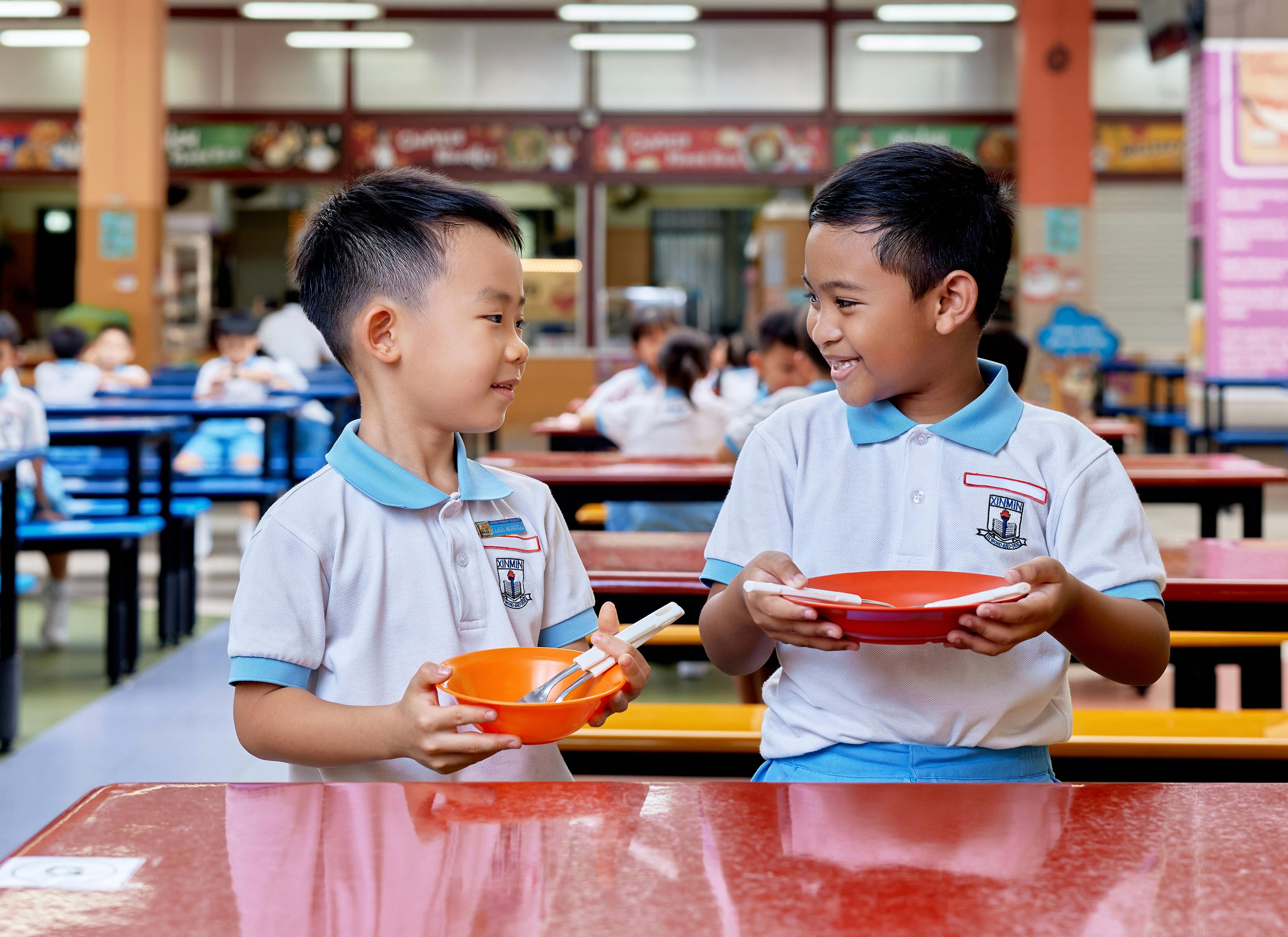 Two schoolboys in uniform smile at each other in a cafeteria, holding bowls with spoons.